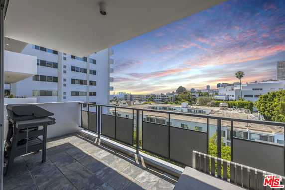 Balcony view overlooking urban surroundings at sunset, with neighboring buildings and a grill on the balcony.