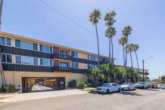 Front view of a multi-story apartment building with a gated entrance and parked cars nearby.