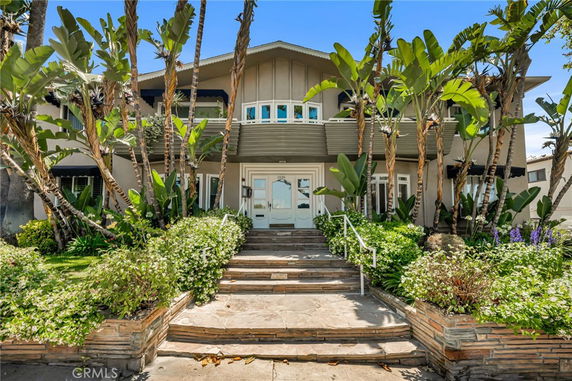 Front view of a two-story house with a staircase leading to the entrance, surrounded by lush vegetation.