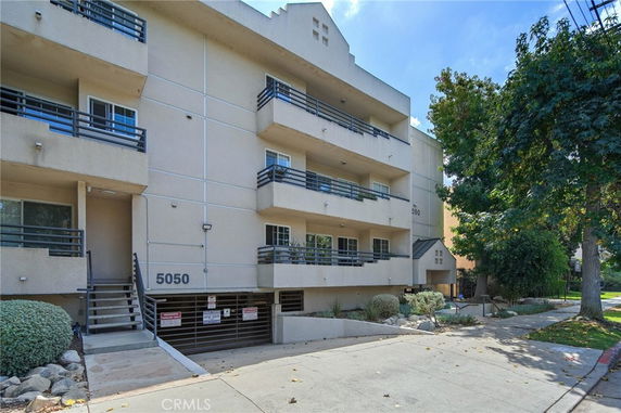 Front view of a multi-story apartment building with balconies and a ground-level parking entrance.