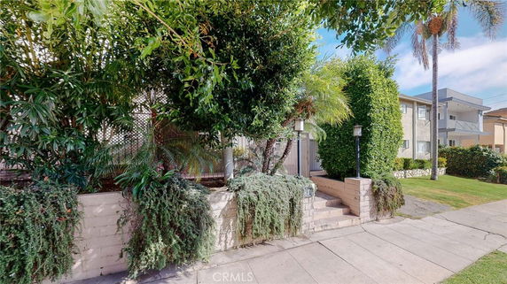 Front view of a multi-story residential building partially obscured by dense greenery and a brick wall.
