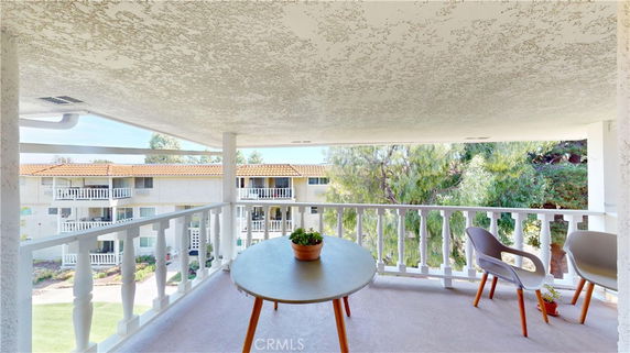 Covered patio area with a table and chairs, offering a view of surrounding buildings and trees.