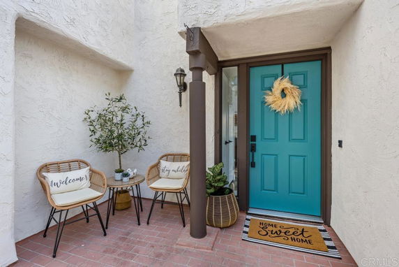 Front entrance of a house with a blue door, decorative wreath, chairs, and a welcome mat on a brick floor.
