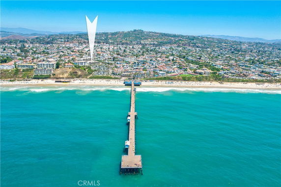 Aerial view of a coastal area with a pier extending into the ocean and a cityscape in the background.