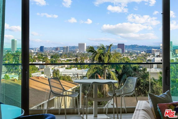 Panoramic view of a cityscape as seen from a building's window, with a balcony furniture setup in the foreground.