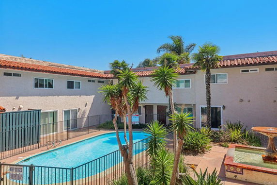 View of a building with an outdoor swimming pool surrounded by palm trees and a fence.