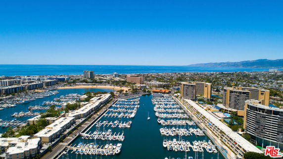 Aerial view of a marina with numerous boats docked, surrounded by residential and commercial buildings, with a coastline and distant mountains in the background.