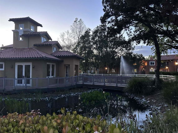 House with multiple roofs and a balcony, adjacent to a pond with a fountain.