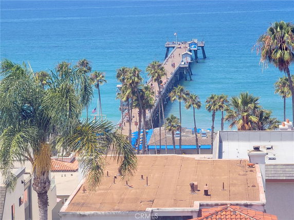 View of a pier extending into the ocean with palm trees and rooftops in the foreground.