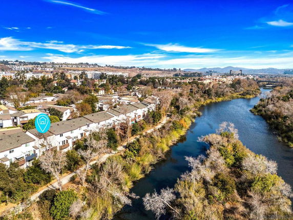 Aerial view showing a residential area by a river with multiple houses, trees, and distant buildings under a clear blue sky.