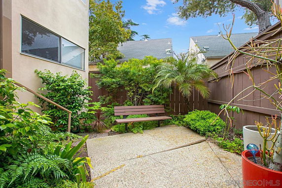 Small outdoor patio with a bench, surrounded by plants and trees, next to a building wall.