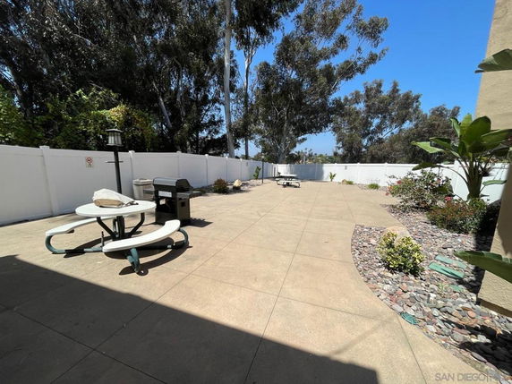 Outdoor area with a circular picnic table, grill, and paved patio surrounded by a white fence and trees.