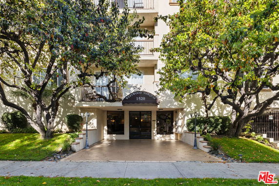 Front view of a multi-story apartment building with trees and a concrete walkway leading to the entrance.