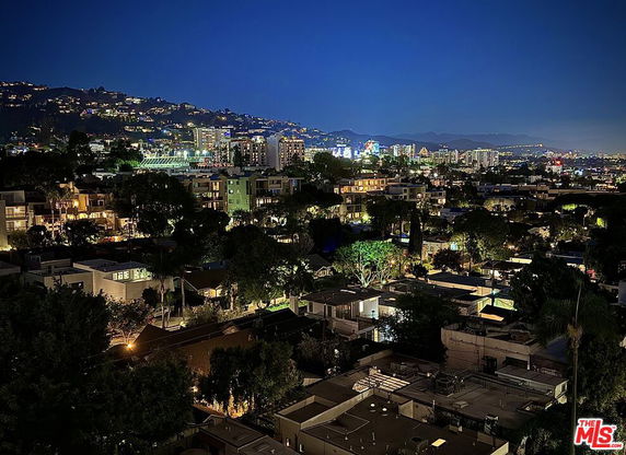 Panoramic night view of cityscape with illuminated buildings and hills in the background.