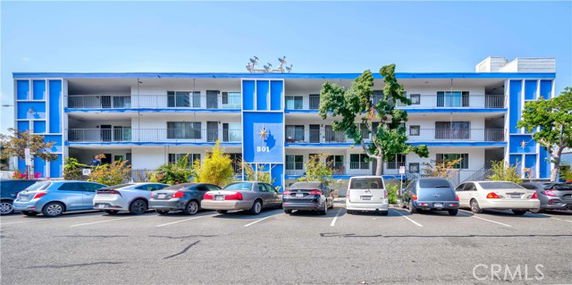 Front view of a three-story blue and white building with balconies and a parking area in front.