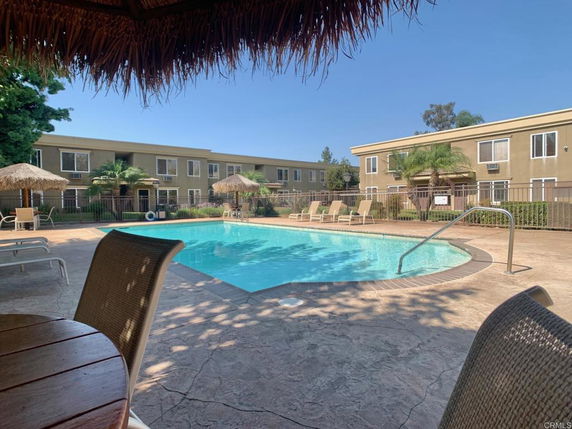 Outdoor swimming pool area with patio chairs and umbrellas, surrounded by a building complex.