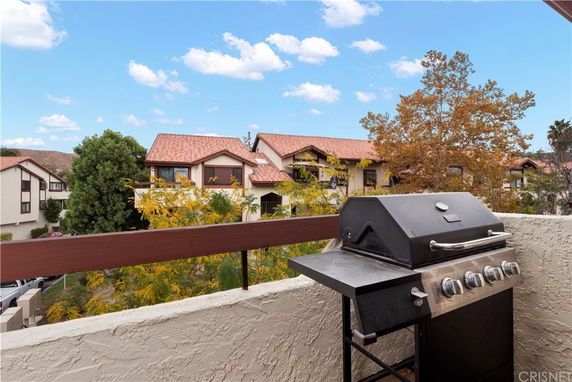 Panoramic view from a balcony with a barbecue grill, showing neighboring houses with reddish-brown roofs and trees.