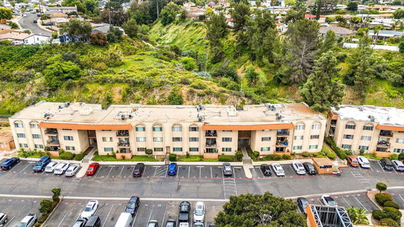 Aerial view of an apartment building with a parking area in front and a lush, green hill behind.