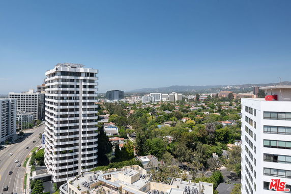 Panoramic view from a high-rise building overlooking other tall buildings and a green residential area with mountains in the distance.