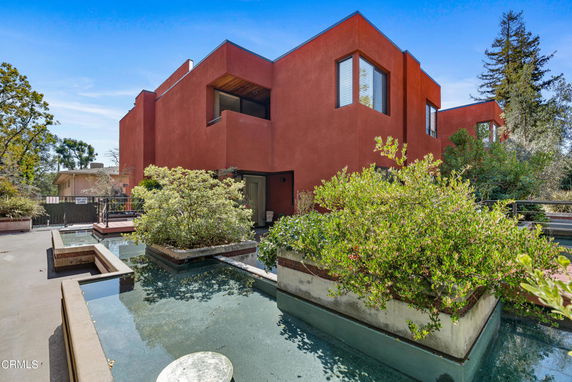 Front view of a modern two-story building with a flat roof and red exterior, surrounded by landscaped plants and water features.