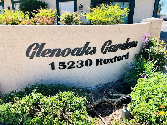 Front view of a building entrance with a wall displaying the name 'Glenoaks Gardens' and an address '15230 Roxford'.