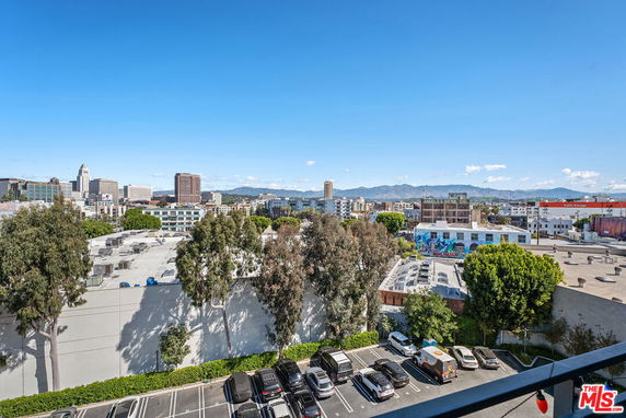 Cityscape view from a higher vantage point overlooking buildings, trees, and parking areas with distant mountains in the background.