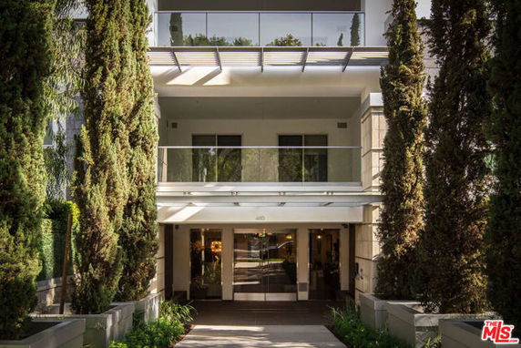 Front view of a building entrance with glass doors and a balcony above, flanked by tall trees.