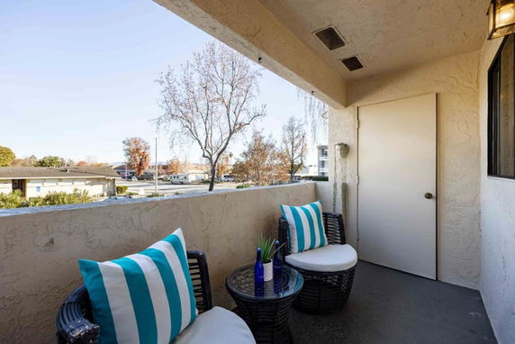 Balcony view from a building showing outdoor seating with chairs and a table, overlooking nearby trees and buildings.