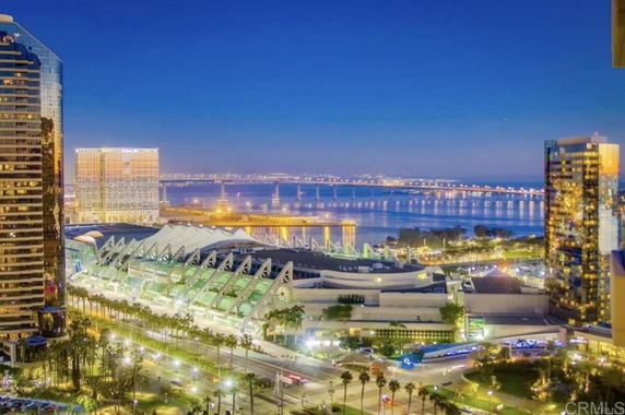 Panoramic view of a waterfront cityscape at dusk with illuminated buildings and a bridge in the background.