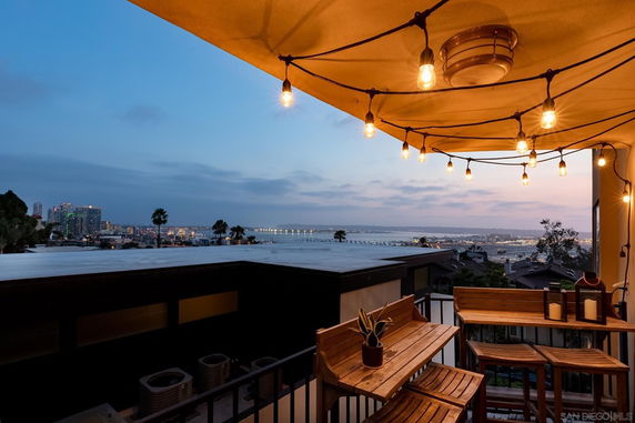 Panoramic view from a rooftop with a canopy and string lights, overlooking a city skyline and body of water at dusk.