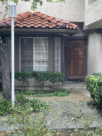 Front view of a house with a wooden door and a small porch area, featuring red tiled roofing and surrounded by shrubs.