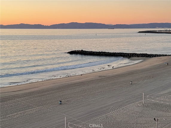 Wide view of a sandy beach with the ocean, breakwaters, and distant hills during sunset.