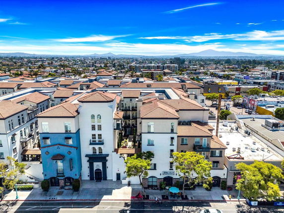 Aerial view of a residential building complex with terracotta roofs, surrounded by urban landscape and distant mountains.