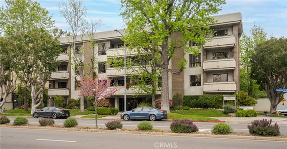 Front view of a four-story apartment building with balconies and surrounding trees.