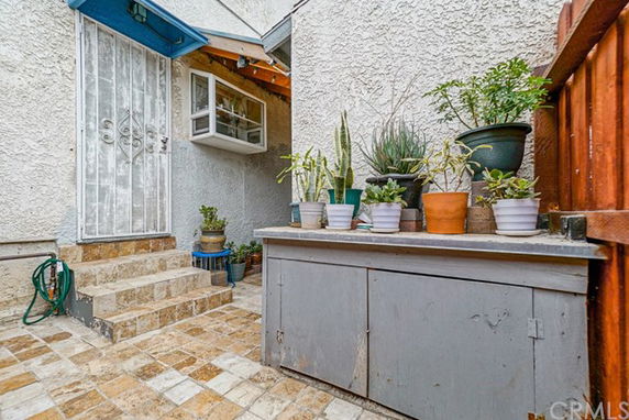 View of an exterior wall with a metal door and steps leading up to it, surrounded by potted plants and tiled flooring.