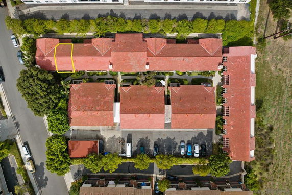 Aerial view of multiple connected buildings with red roofs and a parking area with several parked cars.