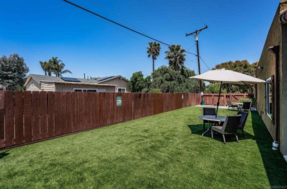 Outdoor seating area with tables, chairs, and umbrellas on a lawn near a fenced boundary.