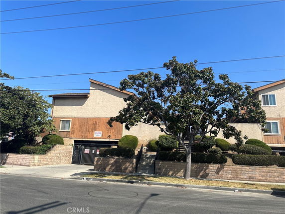 Front view of a multi-story building with a wooden and stucco exterior, surrounded by trees and shrubs.