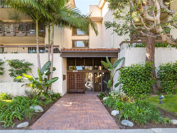 Front entrance of a building with brick walkway and tropical plants.