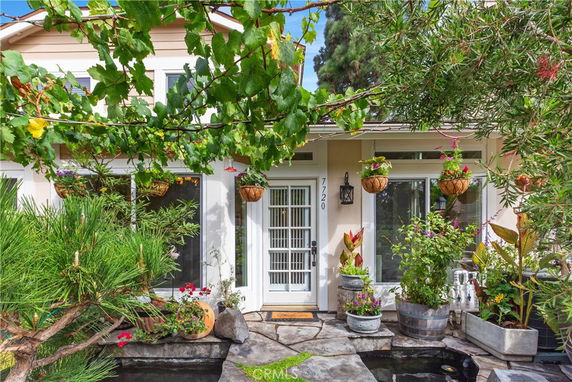 Front view of a house with a white door and large windows, surrounded by plants and hanging baskets.