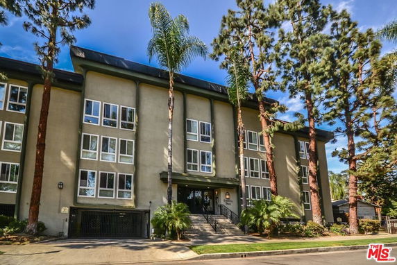 Front view of a multi-story residential building with large windows and a central entrance, surrounded by tall trees.