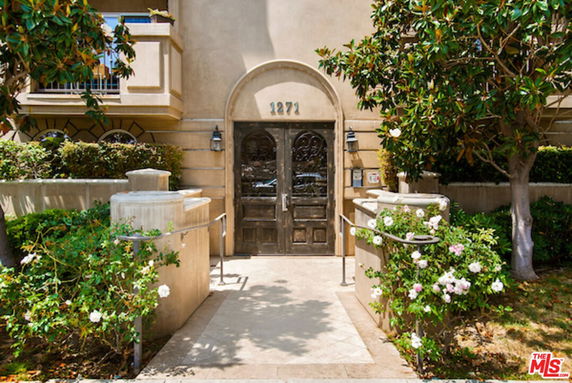 Front view of a building entrance with a double wooden door, arched doorway, and surrounding greenery.