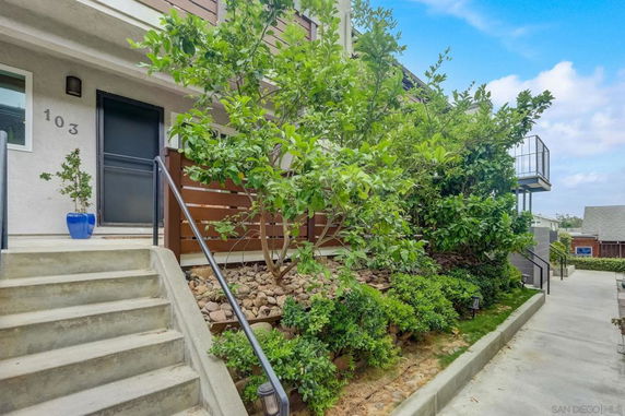 Front view of a building's entrance with stairs leading up to a door marked 103, surrounded by plants and shrubbery.