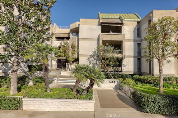 Front view of a multi-story building with a modern design, featuring balconies and a landscaped entrance.