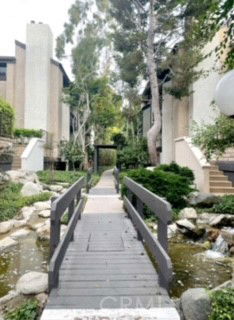 Pathway flanked by modern buildings and trees, with a small bridge over water.
