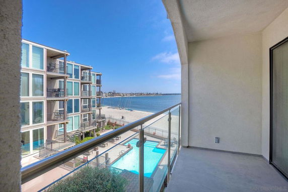 Balcony view overlooking a swimming pool, beach, and distant buildings across the water.