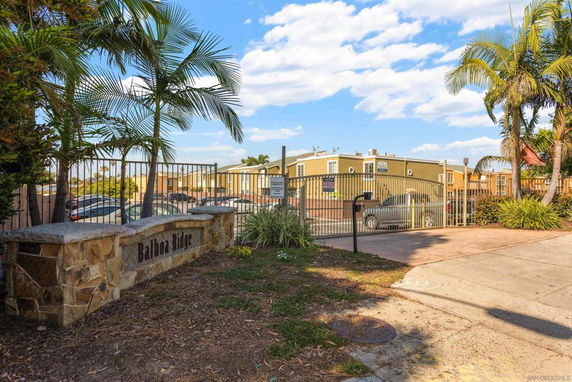 Gated entrance to a residential complex with palm trees and cars visible behind the gate.