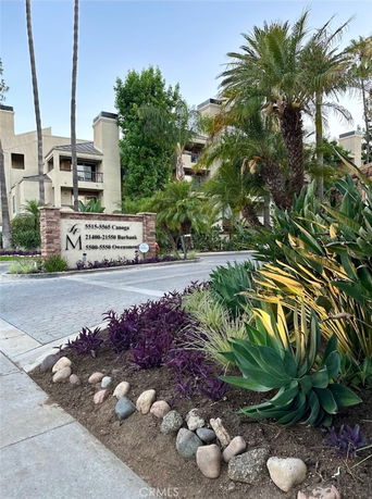 Front view of a multi-story building with balconies and a variety of plants and trees at the entrance.