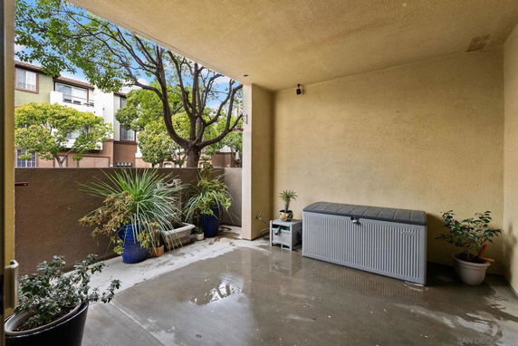 Covered outdoor area with potted plants and a storage box, with trees and buildings visible beyond a low wall.