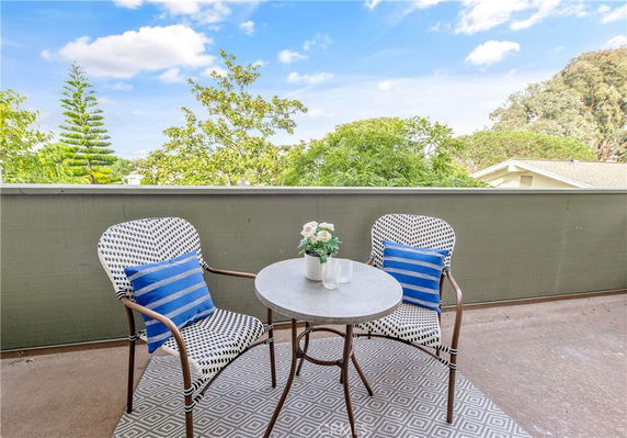 Small outdoor balcony with a round table and two chairs, surrounded by trees and a view of rooftops in the background.